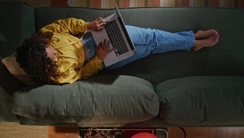 A young woman uses her laptop while sitting on comfortable sofa. A black hispanic girl accessing social media on computer. From above angle of person browsing internet on computer on couch at night